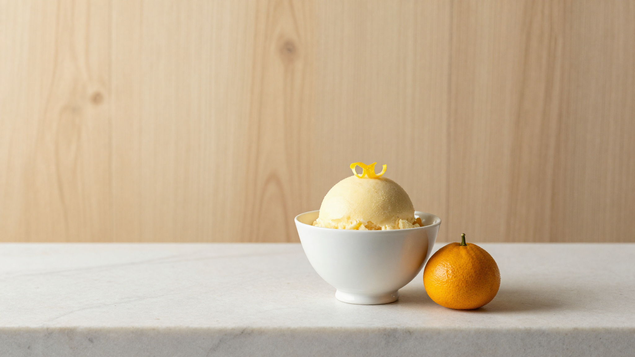 A serving of pale yellow yuzu sorbet in a white ceramic cup garnished with yuzu zest with fresh yuzu fruit beside it